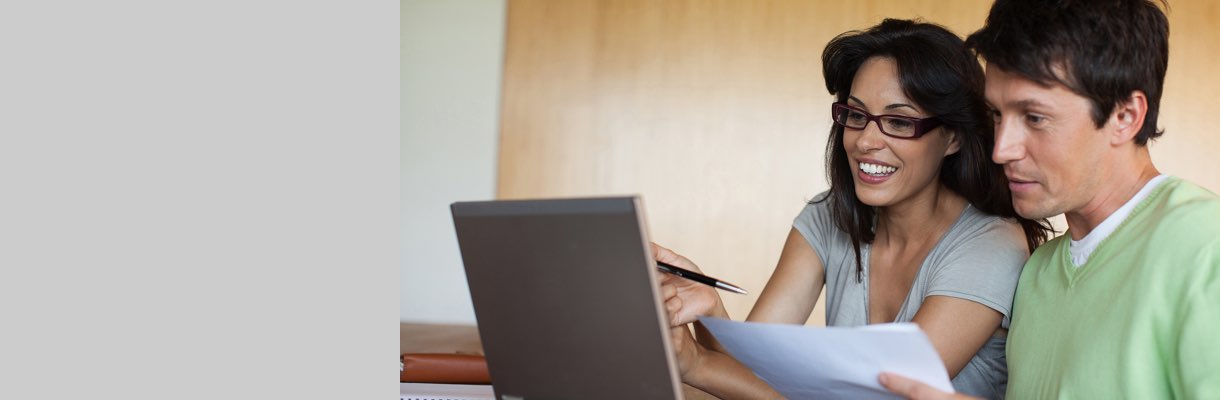 Imagem mostra casal olhando para tela de um computador. A mulher segura uma caneta e o homem segura folhas de papel.