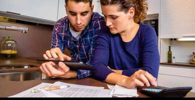 Homem e mulher olham para tablet que ele segura, enquanto mulher faz conta em calculadora sobre a mesa, sobre a qual também estão folhas de papel e caneta