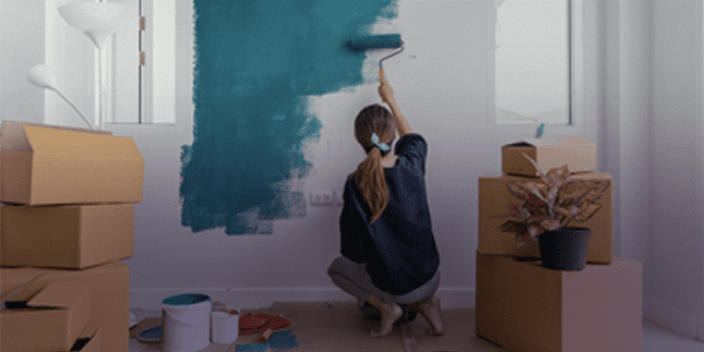 Imagem de mulher ajoelhada pintando parede branca de azul em meio a caixas de papelão